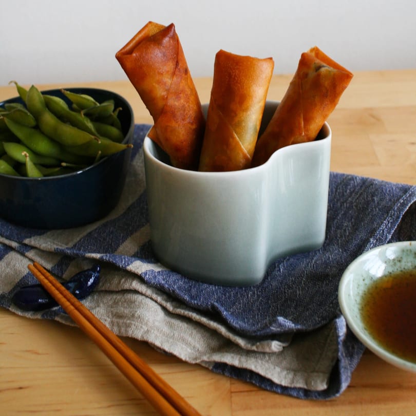 Crispy spring rolls in a white dish, with edamame, chopsticks, and dipping sauce on a wooden table.