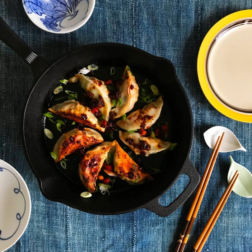 Pan-fried dumplings with chili oil and green onions in a cast iron skillet on a blue tablecloth.