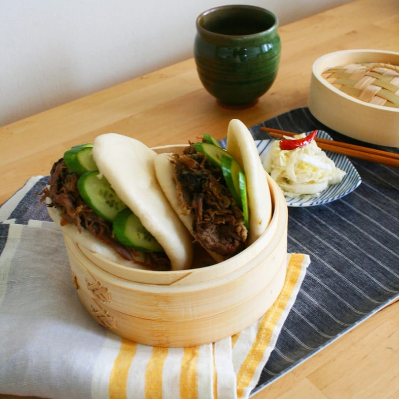 Steamed bao buns with shredded meat and cucumber, served in a bamboo steamer with pickled vegetables.